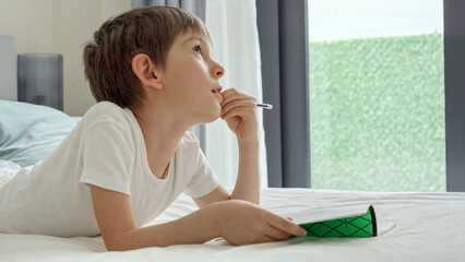 Portrait of smart boy lying in bed at morning and doing homework in notebook. Concept of education, child development and remote schooling at home.