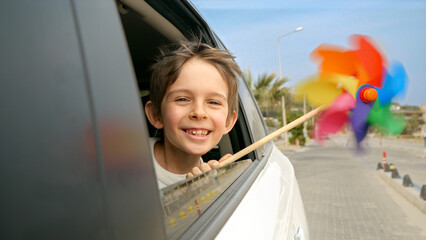 Boy is seen holding a colorful spinning pinwheel as he looks out of the open car window with a smile. Joy of children's travel, journey, tourism, holiday, weekend, and vacation.
