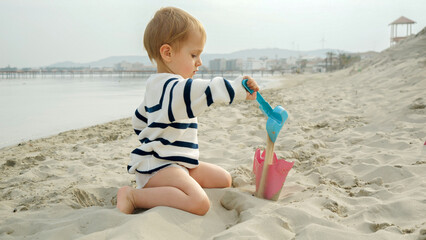 Happy little baby boy enjoying time on the beach and playing with sand using a shovel. Carefree happiness and fun of a family vacation.