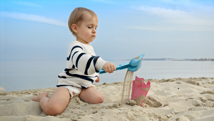 Little boy playing on the sandy beach with toys and using a shovel to dig sand. Happiness and excitement of a family vacation and trip.