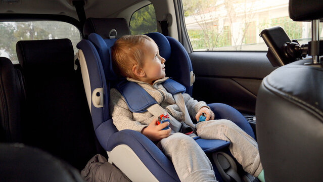 Little baby by sitting in the safety child car seat and looking out of the open window. Concept of kids traveling, baby safety, children going by car.