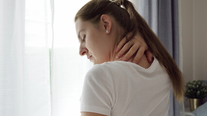 Young woman sitting in bedroom and rubbing or massaging her aching neck and back. Concept of healthcare problems, pain relief and injuries.