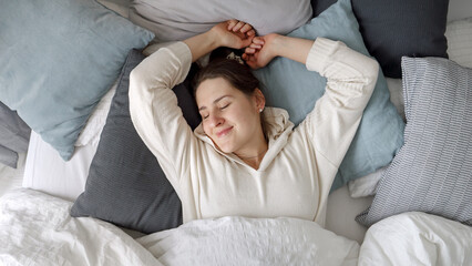Fototapeta na wymiar Relaxed young woman lying on soft pillows and stretching in bed at morning. Concept of comfort, relaxation, healthy sleeping and good start of new day.