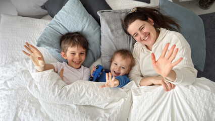 Young happy mother lying in bed with her two sons and waving in camera. Concept of family happiness, relaxing at home, having fun in bed, parent and cheerful kids.
