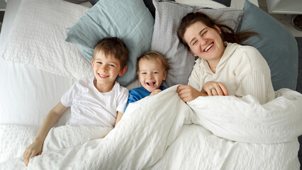 Happy mother with sons lying in bed under blanket and throwing it off smiling at camera. Concept of family happiness, relaxing at home, having fun in bed, parent and cheerful kids
