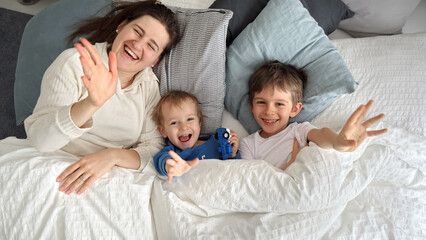 Young happy mother lying in bed with her two sons and waving in camera. Concept of family happiness, relaxing at home, having fun in bed, parent and cheerful kids.