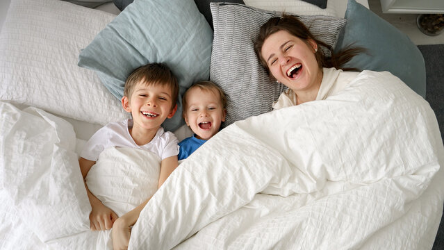 Happy laughing family with two kids hiding under blanket in bed and throwing it off. Concept of family happiness, relaxing at home, having fun in bed, parent and cheerful kids