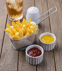 French fries in a basket with salt, ketchup, mustard and a soda glass over wooden table