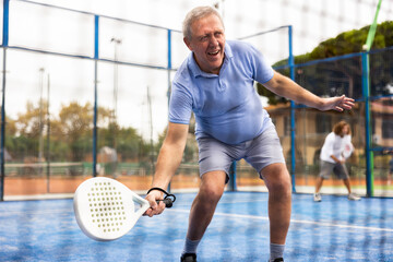 Positive elderly male player serving ball during training padel in court. View through tennis net
