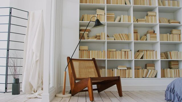 Full zoom shot of stylish expensive wooden chair, retro lamp and large bookcase in library room of spacious modern converted apartment in white shades