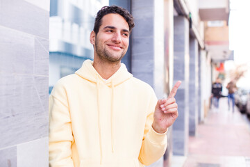 young hispanic man smiling cheerfully, feeling happy and pointing to the side and upwards, showing object in copy space