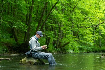 Fisherman choosing trout lures for spinning tackle, sitting on rock in river.