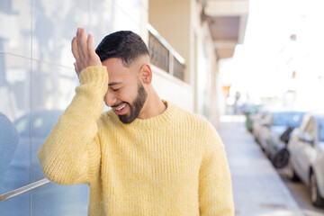 young handsome man laughing and slapping forehead like saying d’oh! I forgot or that was a stupid...