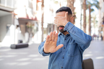 young handsome man covering face with hand and putting other hand up front to stop camera, refusing...