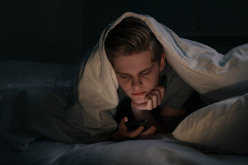 Teenage boy using smartphone under blanket on bed at night. Internet addiction