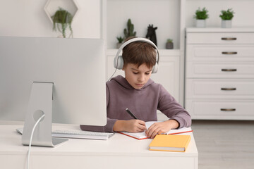 Naklejka premium Boy writing in notepad while using computer and headphones at desk in room. Home workplace