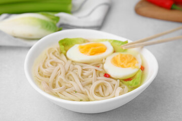 Bowl of delicious rice noodle soup with celery and egg on light grey table, closeup