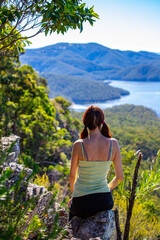 Naklejka premium Beautiful girl sits on a rock shelf enjoying view of the Advancetown lake after successfully climbing the Pages Pinnacle. Hidden gems in Springbrook National Park, Gold Coast, Queensland, Australia