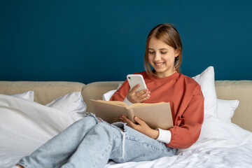 Happy smiling gen Z teen girl relax sitting on bed with book distracted from study receive message...