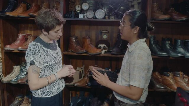 Young Asian seller talking with female customer about old leather boots, consulting her, both standing by shelves with vintage items in antique store