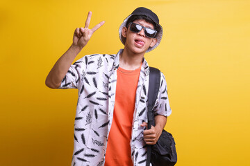 Happy Asian man tourist backpacker having funny expression while show two fingers peace sign symbol isolated on yellow background.  Holiday and travelling promotion concept