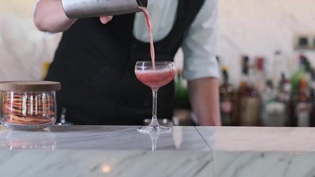 A bartender in an expensive edith hotel pours a prepared pink cocktail from a shaker into a glass and decorates with a slice of lemon