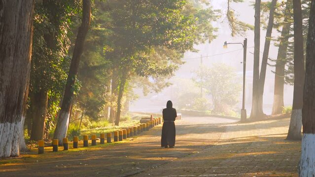 Fraile Franciscano monje cat&oacute;lico cristiano con h&aacute;bito cafe meditando y orando rezando caminando de espaldas en un hermoso camino sendero al amanecer al aire libre en un bosque