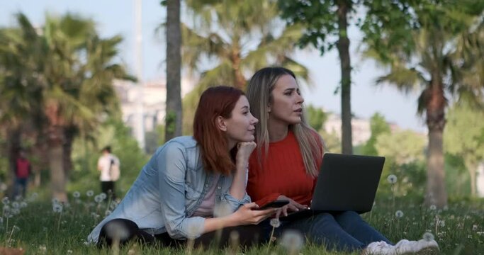 The two young women, sitting in the park, engrossed in their laptops and phones, diligently working on their tasks, become captivated by the scenery and derive immense pleasure from it