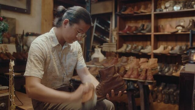 Asian man sitting in vintage store, using bristle brush, polishing old leather boots when restoring them during workday
