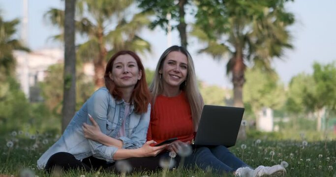 The two young women, sitting in the park, engrossed in their laptops and phones, diligently working on their tasks, become captivated by the scenery and derive immense pleasure from it