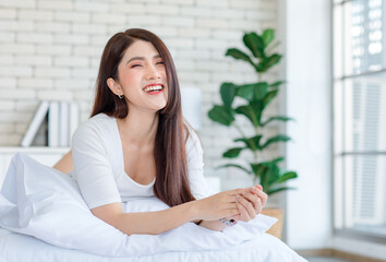 Portrait closeup shot of Millennial Asian beautiful cheerful happy female model in casual outfits with wrist watch laying lying down on white pillow posing smiling look at camera on bed in bedroom