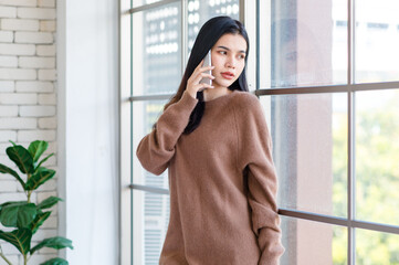 Millennial Asian beautiful cheerful happy female model in casual big brown long sleeve warm sweater outfit standing smiling leaning on glass windows posing holding smartphone waking up in morning