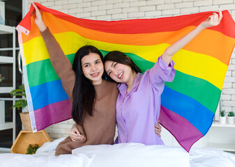 Millennial two Asian beautiful pretty female lesbian girlfriends lover in casual outfits sitting posing on bed in bedroom smiling together holding rainbow equality freedom LGBTQ gay proud pride flag