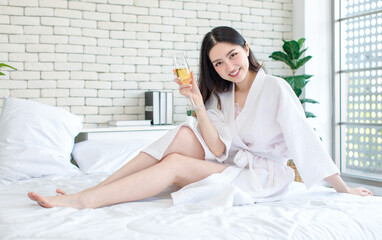 Millennial Asian cheerful happy beautiful female model in casual white clean bathrobe outfit sitting posing smiling holding sparkling champagne tall glass cheer toasting party alone on bed in bedroom