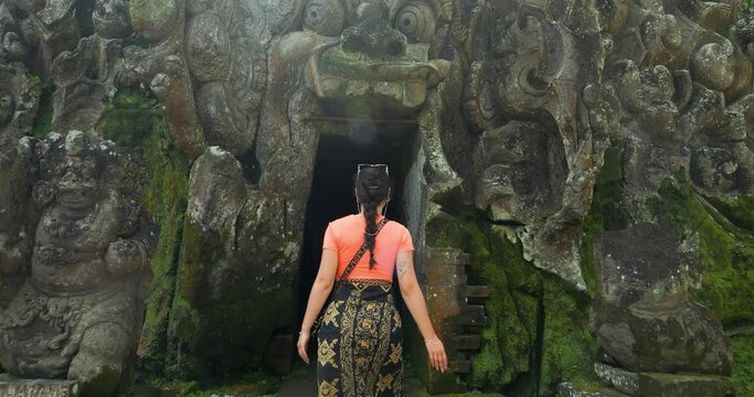 Slow motion dolly shot of a young tourist dressed in a sarong on her trip visiting the Goa Gajah temple in bali while entering the Goa Gajah elephant cave in ubud indonesia