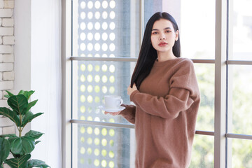 Millennial Asian beautiful cheerful happy female model in casual big brown long sleeve warm sweater outfit standing smiling leaning on glass windows posing holding hot coffee cup waking up in morning