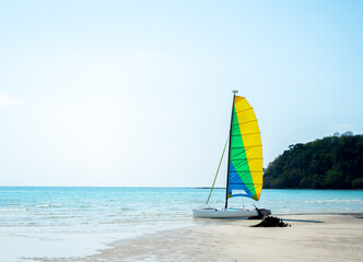 An empty white sailboat with yellow, blue and green pattern sails looks outstanding moored on the sandy beach against the background of the sea, island and blue clear open sky on a sunny summer day.