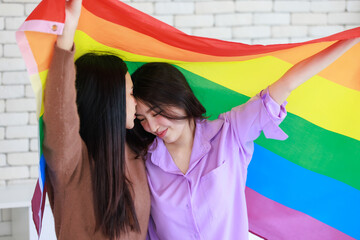 Millennial two Asian beautiful pretty female lesbian girlfriends lover in casual outfits sitting posing on bed in bedroom smiling together holding rainbow equality freedom LGBTQ gay proud pride flag