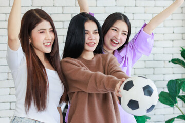 Millennial three Asian beautiful cheerful female girlfriends in casual outfit standing smiling holding football and hands fists up celebrating cheering sport soccer team winning cup victory on TV