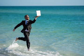 Freelance work, online business. Summer business. Businessman in suit hold laptop and run on sea. Funny businessman in a business suit with laptop standing in water. Remote business.