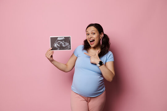 Overjoyed pregnant woman sharing positive emotions looking at camera, pointing aside the ultrasound scan image of her future baby, isolated over pink background, dressed in blue t-shirt and pink pants