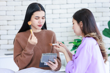Millennial three Asian cheerful happy beautiful female girlfriends in casual outfits sitting resting relaxing holding mirror on bed in bedroom using make up cosmetics lipstick brushing together