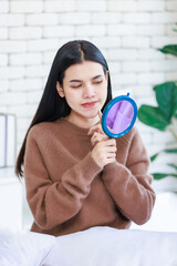 Millennial Asian young beautiful cheerful happy female model in casual long sleeve sweater shirt outfit sitting smiling looking reflection from handheld mirror on bed