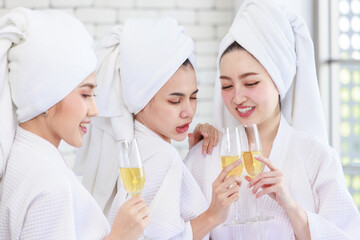 Obraz na płótnie Canvas Three Asian cheerful happy beautiful female girlfriends in casual bathrobe and towel outfit standing smiling holding sparkling champagne tall glass cheers toasting together in spa massage studio