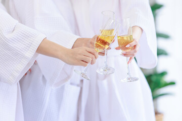 Closeup shot of three tall glasses of sparkling champagne cheering toasting drinking by unrecognizable unknown women in white clean bathrobes celebrating relaxing resting in spa massage studio resort