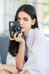 Closeup shot of Millennial Asian beautiful confident female model in white clean bathrobe sitting smiling posing holding mirrorless DSLR camera ready for work with photographer in photography studio