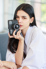 Closeup shot of Millennial Asian beautiful confident female model in white clean bathrobe sitting smiling posing holding mirrorless DSLR camera ready for work with photographer in photography studio