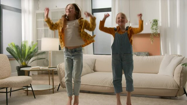 Two active funny little caucasian kids girls having fun moving play at home cheerful joyful children sisters friends playing together jumping dancing listening to music jump enjoy dance in living room