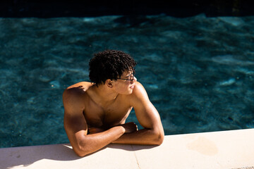 High angle view of biracial shirtless young man in sunglasses standing in pool at poolside