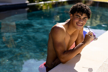 Portrait of biracial shirtless young man holding sunglasses smiling and leaning at poolside